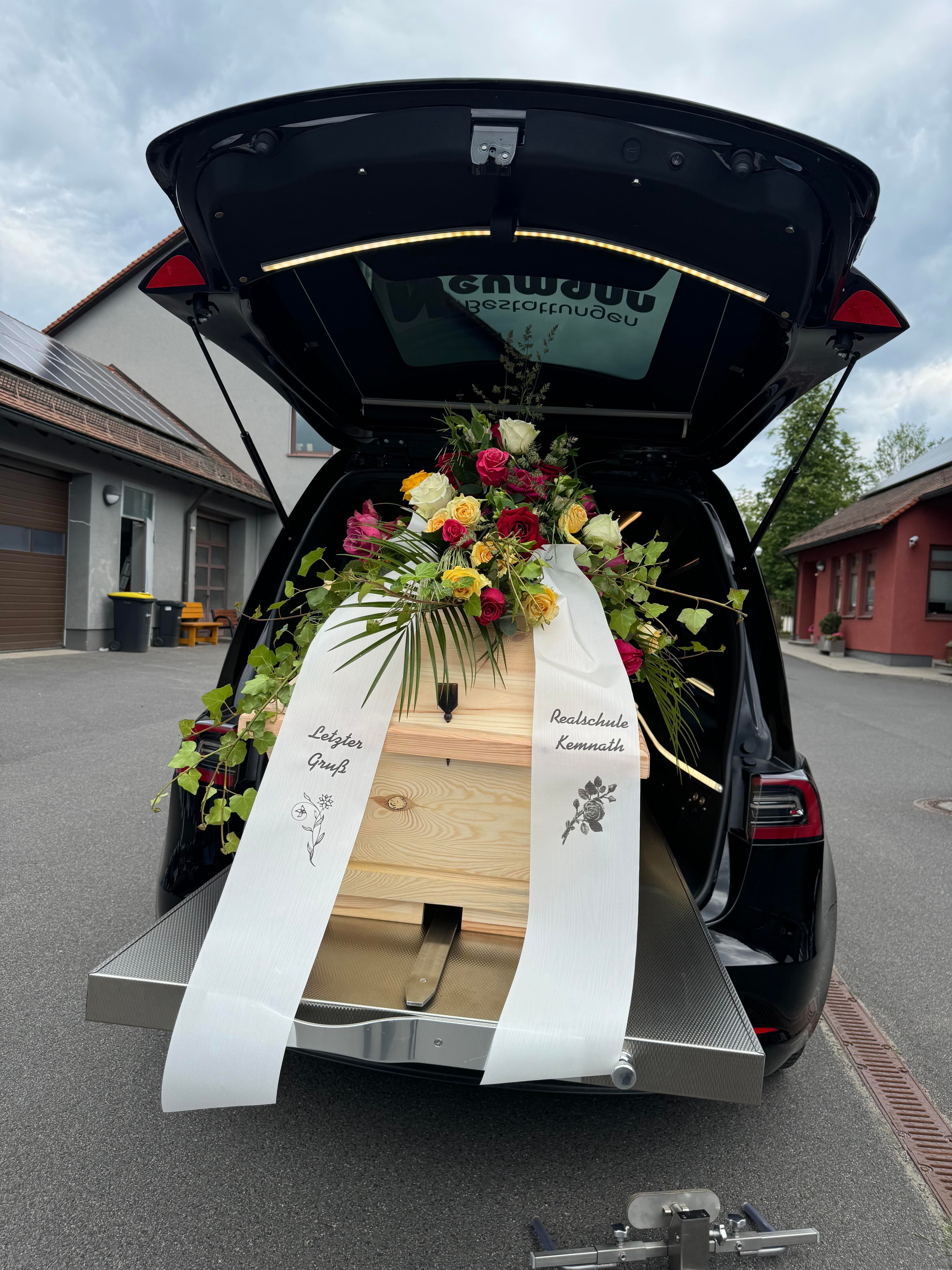 Sarg mit Blumenschmuck im geöffneten Leichenwagen