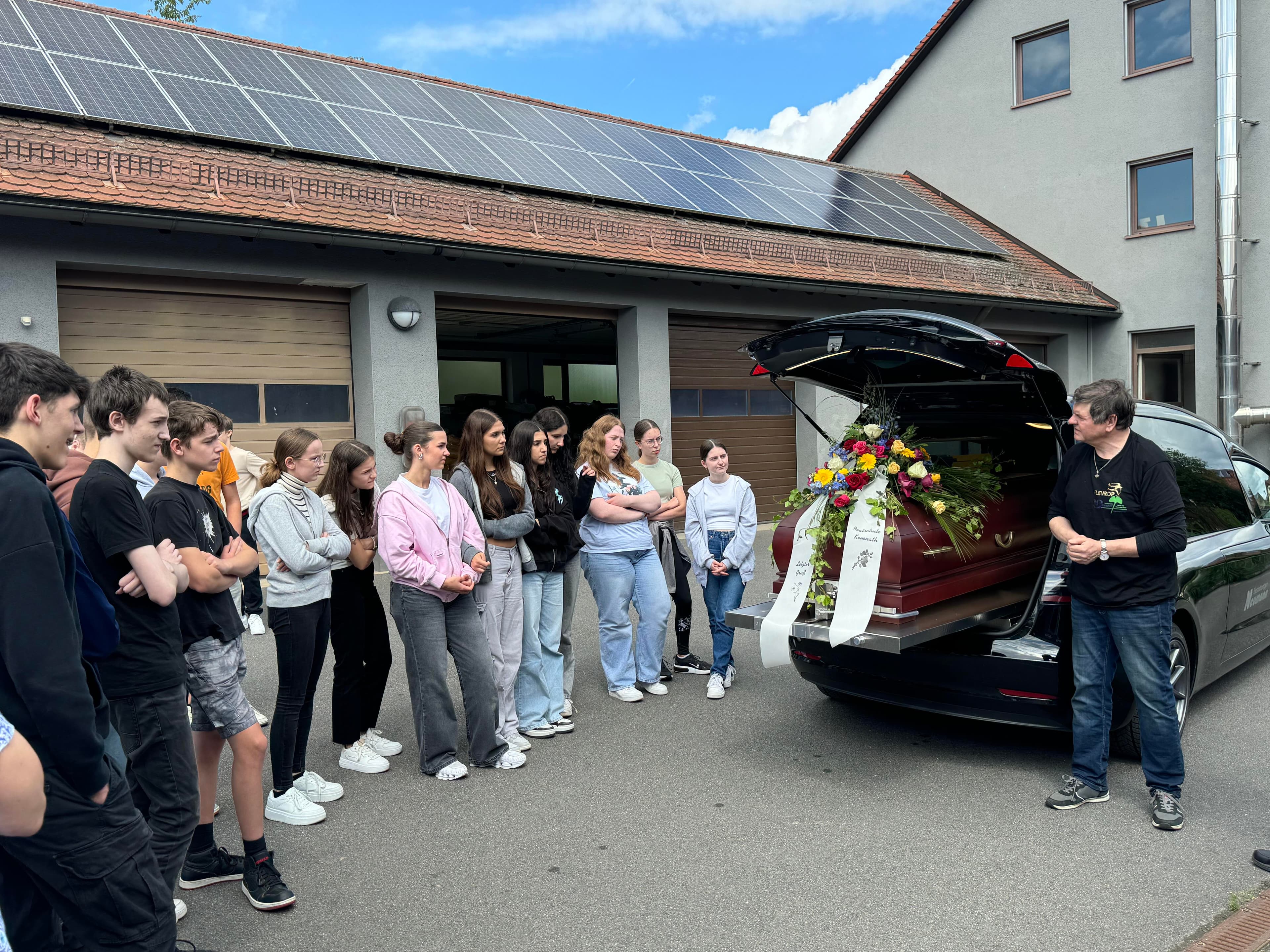 Schulklasse steht vor Leichenwagen mit Blumenschmuck